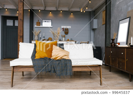 Stylish and Scandinavian interior of the living room of a modern apartment with a white sofa, pillows, a blanket, a kitchen in the background. Modern home decor. 99645837