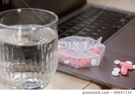 Organizer weekly shots with glass of water on workplace background and laptop Closeup of medical pill box with doses of tablets for daily take medicine with white pink drugs and capsules. Health care Organizer weekly shots with glass of water on workplace background and laptop Closeup of medical pill box with doses of tablets for daily take medicine with white pink drugs and capsules. Health care 99647134