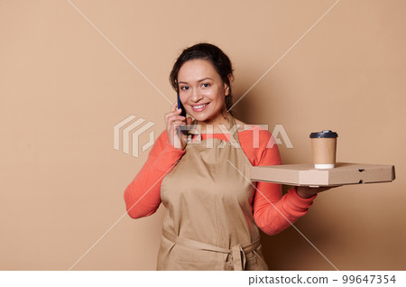 Pretty waitress, female bartender talks on mobile phone, smiling a beautiful toothy smile looking at camera, posing with takeaway hot food in disposable eco containers, isolated on beige background 99647354