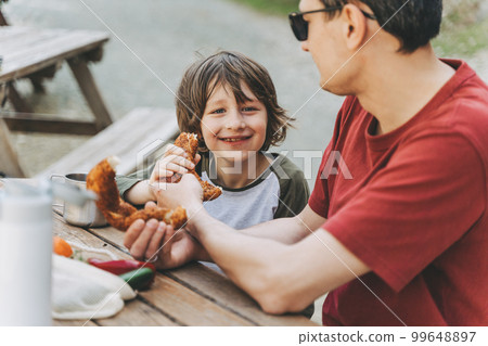 Close-up view of father hugs his school boy son on a family picnic. Child kid and his dad taking a rest and enjoying a picnic while hiking. Boy smiles and bites the bread donut on a picnic 99648897