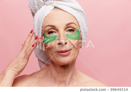 Close-up of beautiful middle-aged woman with towel on head posing with under eye patches, posing over pink studio background. Concept of natural beauty, face skin care, cosmetics 99649314