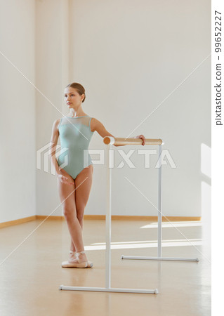 Beautiful teen girl, ballet dancer training at dance school, standing at position near ballet barre on daytime with sunlight. Concept of classical ballet, art, choreography 99652227