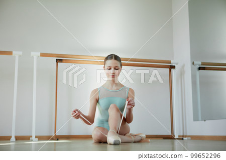 Attractive. Tender young girl, ballerina sitting on floor and putting on pointe shoes. Training in ballet school, indoors. Concept of classical ballet, art, choreography Attractive. Tender young girl, ballerina sitting on floor and putting on pointe shoes. Training in ballet school, indoors. Concept of classical ballet, art, choreography 99652296