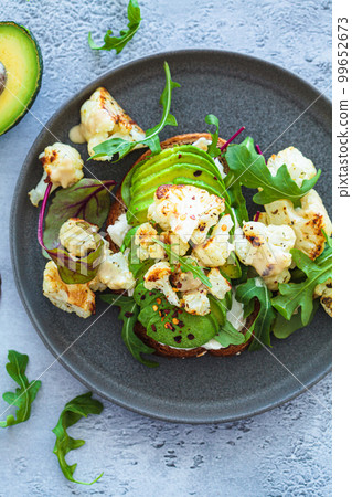 Vegan toast with avocado, baked cauliflower, lettuce and tahini on dark plate. Healthy plant based organic diet. 99652673