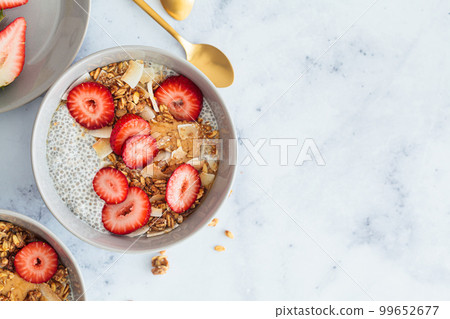 Chia pudding with homemade coconut granola, peanut butter and strawberries in a gray bowl, marble background. Healthy diet, detox, summer recipe. 99652677
