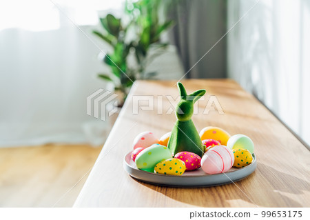 Easter home decor. Green bunny rabbit figurine and colored easter chocolate eggs on plate on wooden console with green home plants and sunlight and shadows on white wall. Selective focus. Copy space. Easter home decor. Green bunny rabbit figurine and colored easter chocolate eggs on plate on wooden console with green home plants and sunlight and shadows on white wall. Selective focus. Copy space. 99653175