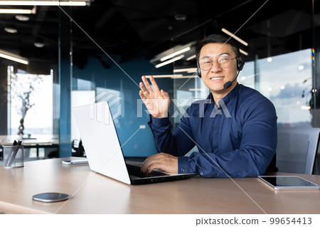 Man at work with headset phone and laptop successful asian smiling and looking at camera at workplace inside office, portrait of successful businessman support worker waving hand greeting gesture Man at work with headset phone and laptop successful asian smiling and looking at camera at workplace inside office, portrait of successful businessman support worker waving hand greeting gesture 99654413