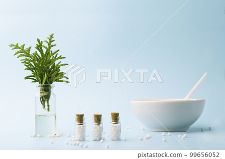 A bottle of essential oil with fresh juniper branches. Homeopathic globules and glass bottles near a white plate. 99656052