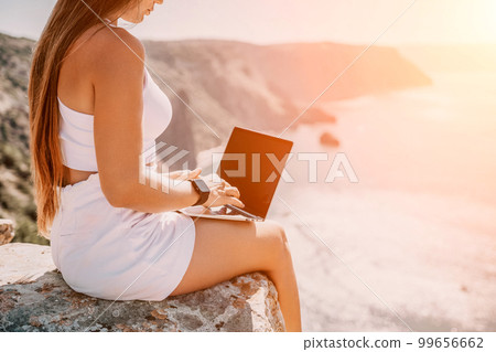 Woman laptop sea. Working remotely on seashore. Happy successful woman female freelancer in straw hat working on laptop by the sea at sunset. Freelance, remote work on vacation 99656662