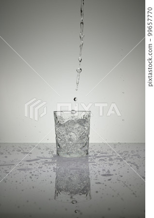 Vertical shot of water pouring into crystal glass with droplets around, on a white shiny table Vertical shot of water pouring into crystal glass with droplets around, on a white shiny table 99657770