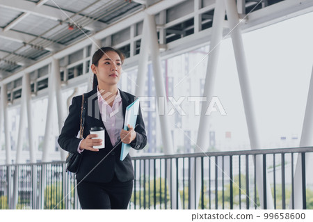 young Asian businesswoman in black suit holding folder and cup of coffee and standing on walkway station. young Asian businesswoman in black suit holding folder and cup of coffee and standing on walkway station. 99658600
