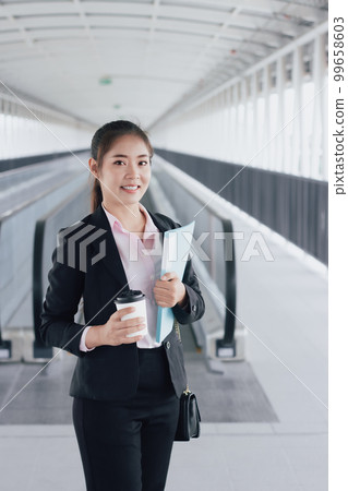 young Asian businesswoman in black suit holding folder and cup of coffee and standing on walkway station. 99658603