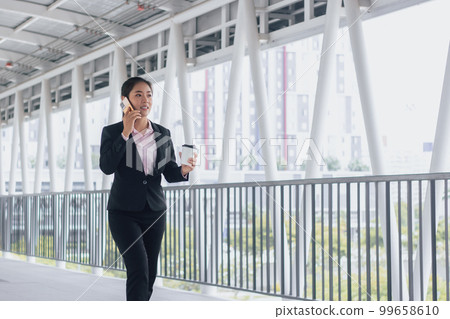 young Asian businesswoman in black suit using phone and holding cup of coffee and standing on elevated walkway station with copy space. 99658610