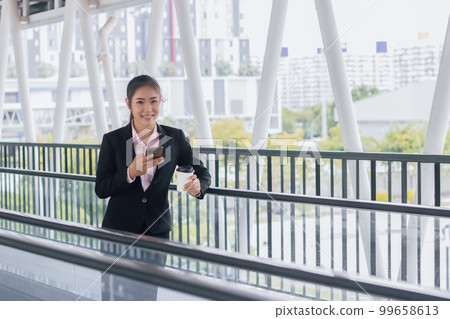 young Asian businesswoman in black suit using phone and holding cup of coffee and standing on elevated walkway station with copy space. young Asian businesswoman in black suit using phone and holding cup of coffee and standing on elevated walkway station with copy space. 99658613