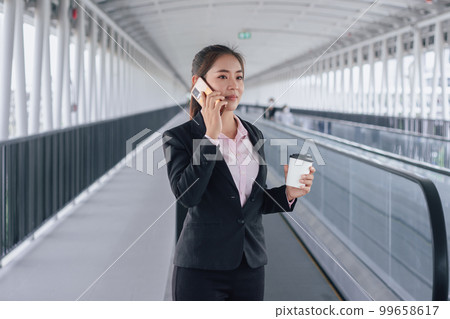 young Asian businesswoman in black suit using phone and holding cup of coffee and standing on elevated walkway station with copy space. 99658617