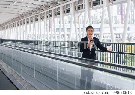 young Asian businesswoman in black suit using phone and holding cup of coffee and standing on elevated walkway station with copy space. 99658618