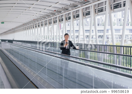 young Asian businesswoman in black suit using phone and holding cup of coffee and standing on elevated walkway station with copy space. young Asian businesswoman in black suit using phone and holding cup of coffee and standing on elevated walkway station with copy space. 99658619