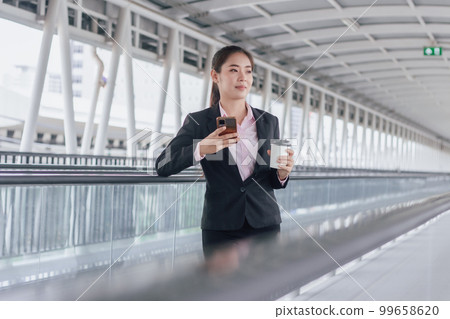 young Asian businesswoman in black suit using phone and holding cup of coffee and standing on elevated walkway station with copy space. 99658620
