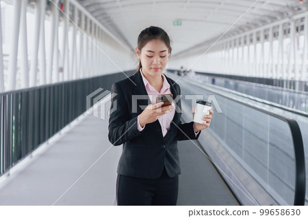young Asian businesswoman in black suit using phone and holding cup of coffee and standing on elevated walkway station with copy space. young Asian businesswoman in black suit using phone and holding cup of coffee and standing on elevated walkway station with copy space. 99658630