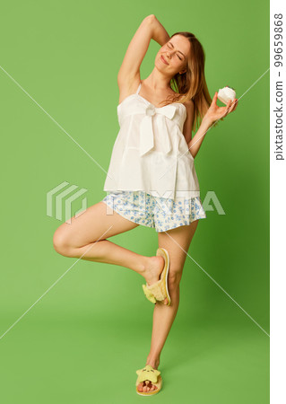 Beautiful cheerful woman wearing comfortable pyjamas, homewear eats sweet cake and rejoices isolated on green background. 99659868