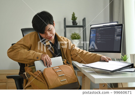Smiling asian male programmer talking on mobile phone and putting his stuff brief case into bag at workplace 99660671