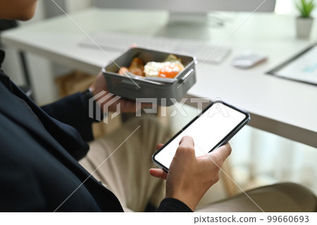 Cropped shot of male employee holding plastic container and using smart phone at his working desk Cropped shot of male employee holding plastic container and using smart phone at his working desk 99660693