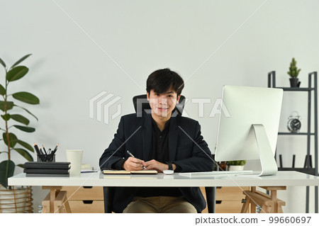 Portrait of young start up businessman in elegant suit sitting at his workplace in the modern office and smiling to camera Portrait of young start up businessman in elegant suit sitting at his workplace in the modern office and smiling to camera 99660697