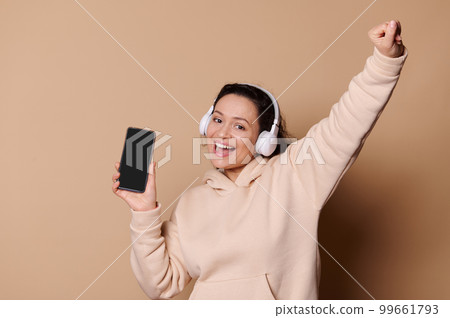 Smiling pretty woman with headphones having fun, holding smartphone with black with free ad space for text or mobile app. Pretty woman listening to music, enjoying playlist, beige isolated background Smiling pretty woman with headphones having fun, holding smartphone with black with free ad space for text or mobile app. Pretty woman listening to music, enjoying playlist, beige isolated background 99661793
