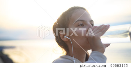 Young beautiful sportive girl listening to music and drinking water at sunrise by the lake. Young beautiful sportive girl listening to music and drinking water at sunrise by the lake. 99663155