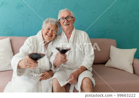 Happy senior couple sitting together in bathrobe on sofa with glass of wine, having nice time at home. Happy senior couple sitting together in bathrobe on sofa with glass of wine, having nice time at home. 99663222