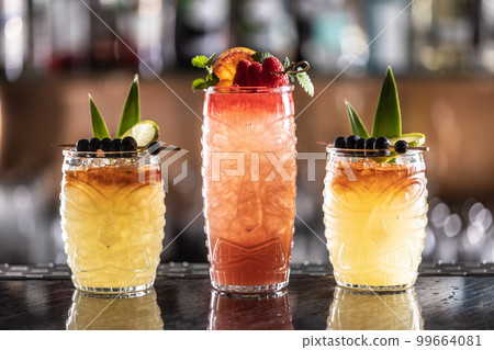 Mai tai cocktails in hawaiian glasses garnished with pineapple, blueberries and strawberries on bar counter. 99664081