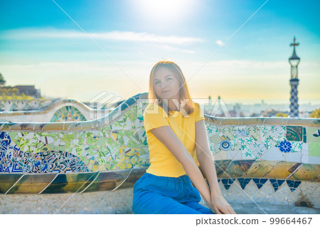 Positive young female tourist sitting on stone bench and checking photos while resting during vacation at Park Guell Positive young female tourist sitting on stone bench and checking photos while resting during vacation at Park Guell 99664467