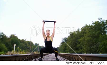 Beautiful sexy athletic young blond woman in black leggings performing exercises with a heavy weight plate, On the railway, on rails, sleepers, in the summer. Slow motion 99665554