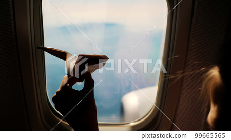 close-up. Silhouette of a child's hand with small paper plane against the background of airplane window. Child sitting by aircraft window and playing with little paper plane. during flight on airplane 99665565