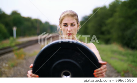 Beautiful sexy athletic young blond woman in black leggings performing exercises with a heavy weight plate, On the railway, on rails, in the summer. Slow motion. portrait Beautiful sexy athletic young blond woman in black leggings performing exercises with a heavy weight plate, On the railway, on rails, in the summer. Slow motion. portrait 99665590