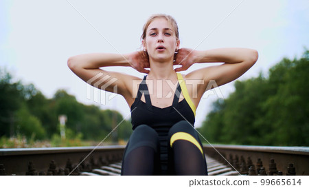 Beautiful sexy athletic young blond woman in black leggings performing exercises to make strong press, abdominal muscles. Sits on sleepers, on railway, in summer, Slowmotion Beautiful sexy athletic young blond woman in black leggings performing exercises to make strong press, abdominal muscles. Sits on sleepers, on railway, in summer, Slowmotion 99665614