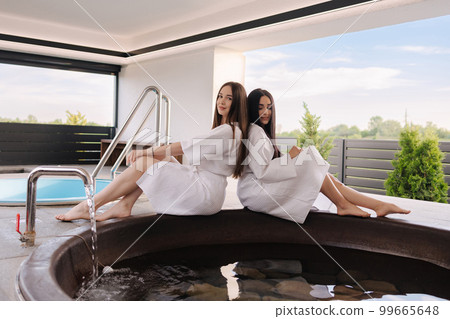 Two beautiful woman in white bathrobe sits on hot tube outdoors. Sexy lady on luxury resort  99665648