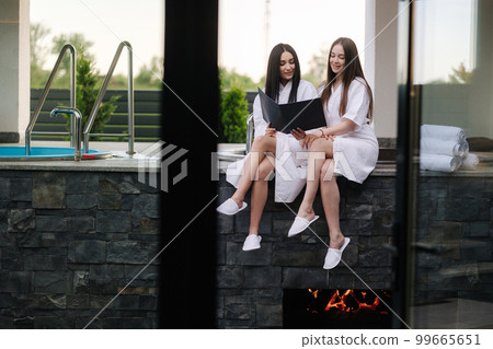 Two beautiful woman in white bathrobe sits in front of hot tube outdoors. Sexy lady on luxury resort  99665651