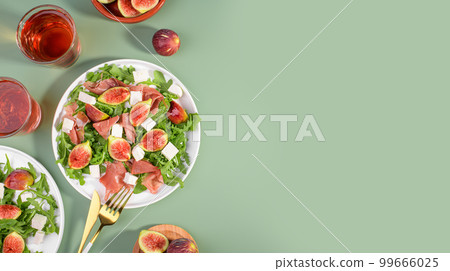Autumn figs salad with arugula, feta, raw ham in white plate on green background served with rose wine, top view 99666025