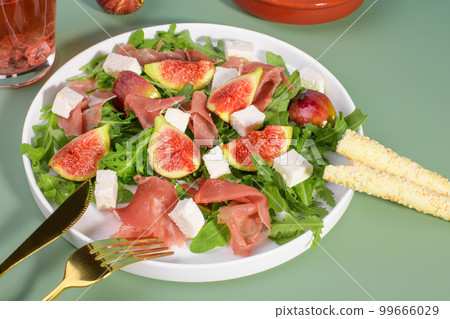 Autumn figs salad with arugula, feta, raw ham in white plate on green background served with rose wine and grissini. 99666029