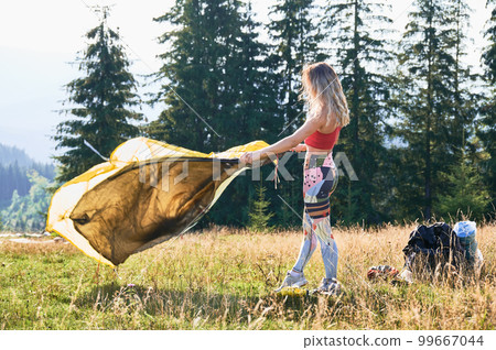 Woman tourist camping in mountains. Female tourist wearing sportswear, putting tent, making campsite. Concept of tourism and beauty of nature. 99667044