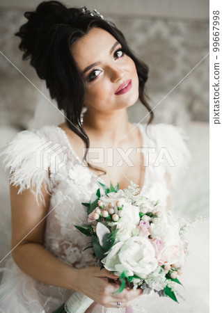 Beautiful bride wearing fashion wedding dress with feathers with luxury delight make-up and hairstyle, studio indoor photo shoot Beautiful bride wearing fashion wedding dress with feathers with luxury delight make-up and hairstyle, studio indoor photo shoot 99667998