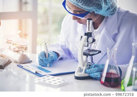 Woman scientist hand write note information in medical lab look at science microscope medical test, research biology chemistry. Asian Females technician laboratory analyzing blood test pharmaceutical 99672358