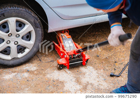 Male man jacked up blue Hyundai car with red jack outside. 99675290