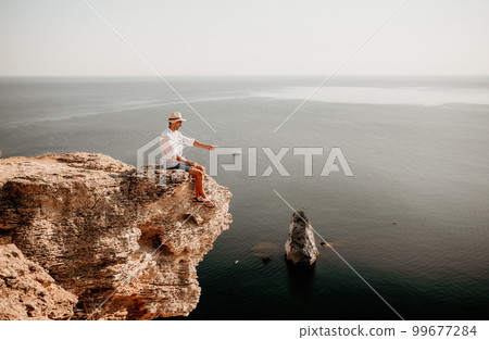 Digital nomad, man in the hat, a businessman with a laptop sits on the rocks by the sea during sunset, makes a business transaction online from a distance. Remote work on vacation. 99677284