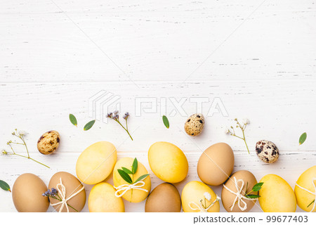 Festive Easter background. Yellow and brown Easter eggs with flowers on a white wooden table. Card with a place for text. Top view. Festive Easter background. Yellow and brown Easter eggs with flowers on a white wooden table. Card with a place for text. Top view. 99677403