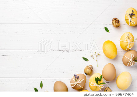 Festive Easter background. Yellow and brown Easter eggs with flowers on a white wooden table. Card with a place for text. Top view. Festive Easter background. Yellow and brown Easter eggs with flowers on a white wooden table. Card with a place for text. Top view. 99677404
