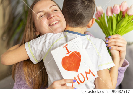 Child son congratulates his mother and gives her postcard and bouquet of tulips 99682517