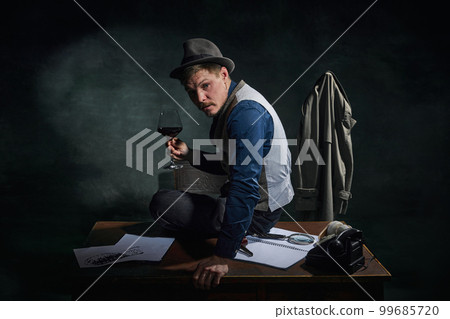 Professional detective in fedora hat and classical clothes sitting on table with glass of red wine over dark green vintage background 99685720