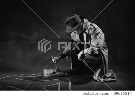 Black and white, noir photography. Man, detective in hat and trench coat looking on human drawn silhouette on floor and picking handkerchief with blood spots 99685814